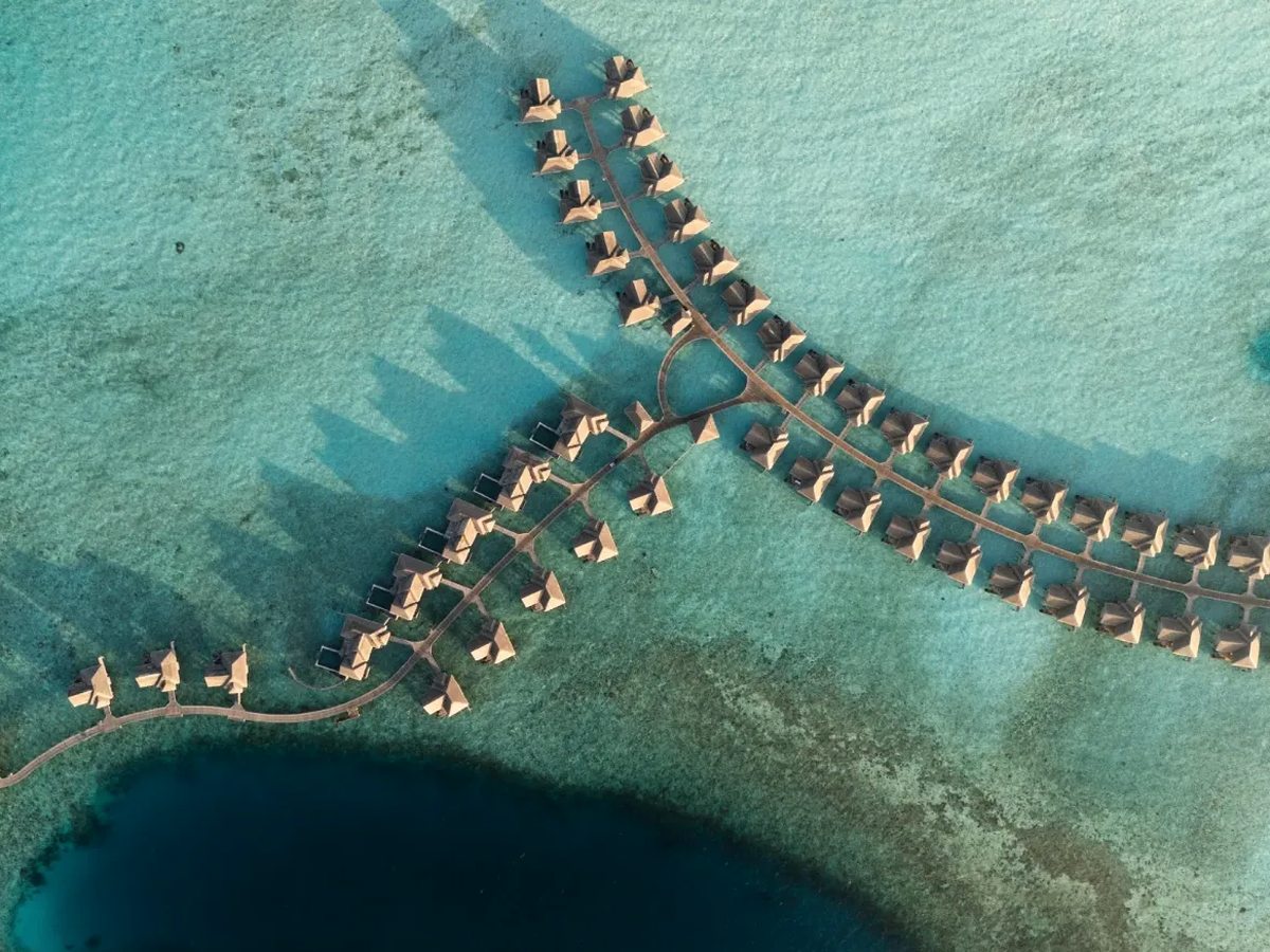 Aerial view of two curving arms of thatched overwater villas standing over pale turquoise water, with a band of deep blue water below them.