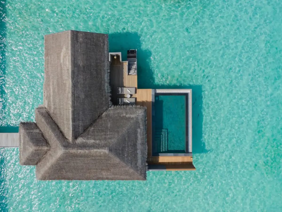 Overhead view of a thatched overwater villa on turquoise water, with a rectangular pool and sun deck on one side.