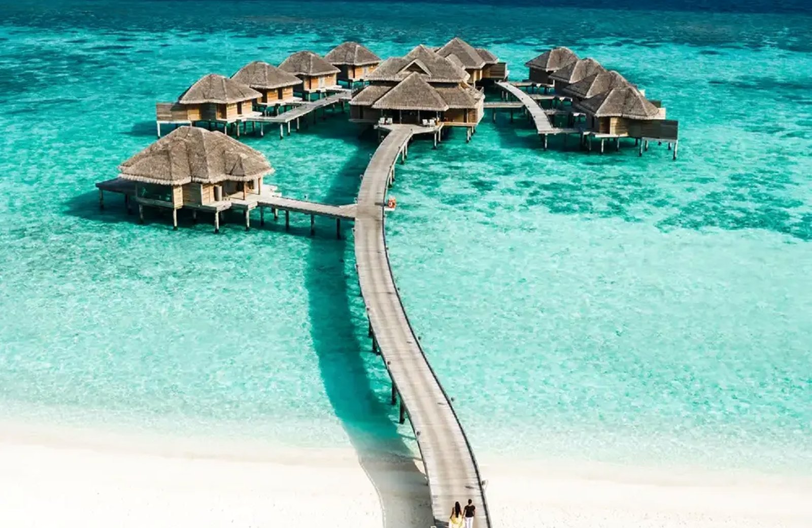 Thatched overwater villas on stilts in a turquoise lagoon, joined by a curving wooden boardwalk that runs back to a white sand beach where two people are walking.
