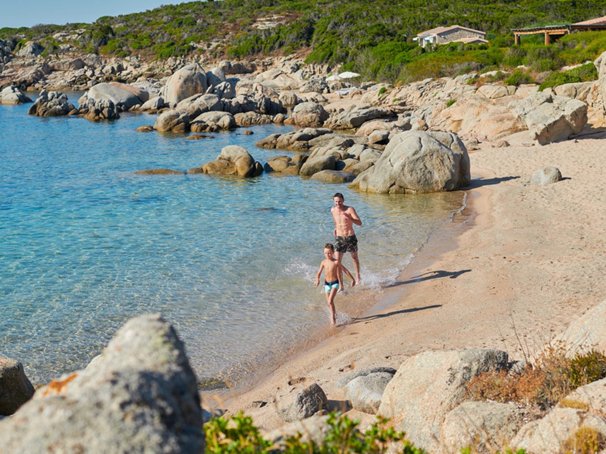 A man and a boy running out of the shallow water onto the sand of a small cove enclosed by large granite boulders, with scrub and a low building on the slope behind.