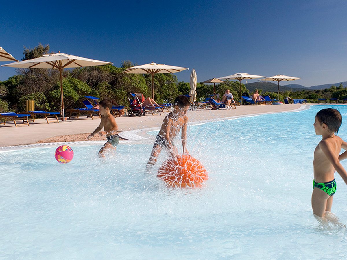 Children playing with an orange and a pink ball in a wide, shallow pool, with cream parasols, blue sun loungers and scrub behind.