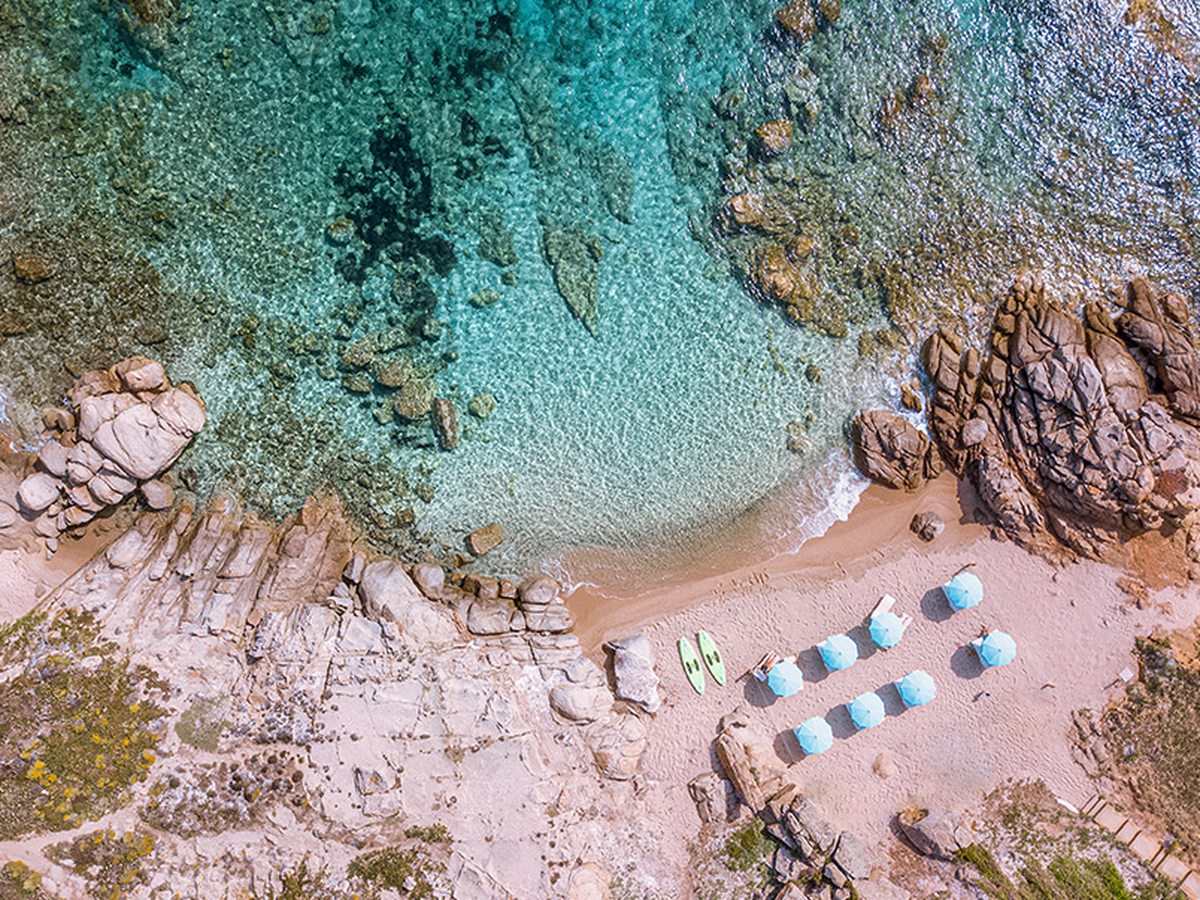Aerial view of a small sandy cove with rows of blue and white parasols and sunbeds on the sand, granite rocks at each end and clear turquoise water.