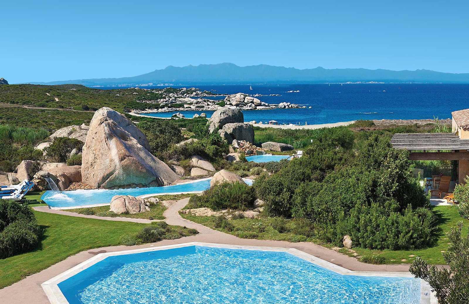 Two of the resort's pools set among granite boulders and lawns, with water falling between them, and a sandy beach and deep blue sea beyond.