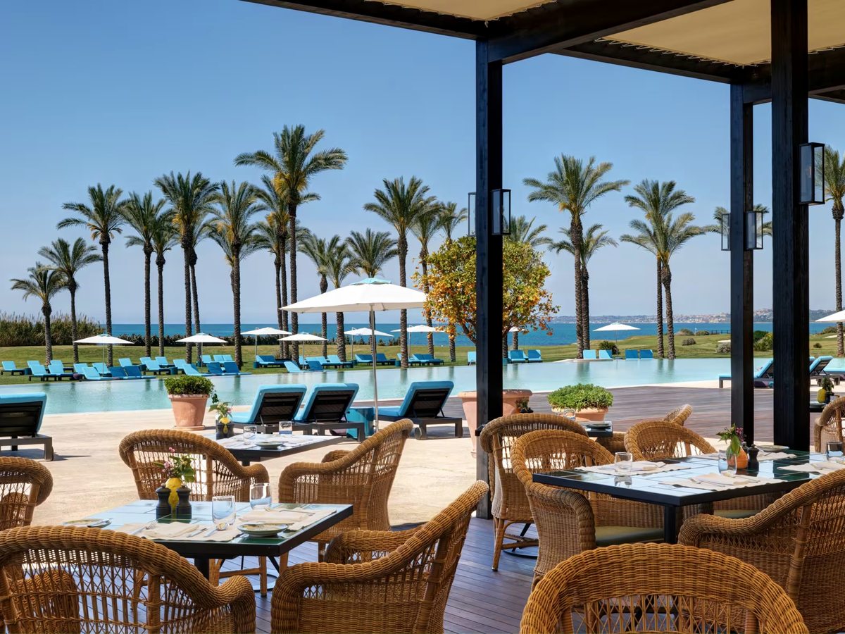 Wicker chairs and laid tables on a timber dining terrace looking across a long infinity pool lined with blue sun loungers and tall palms to the sea.