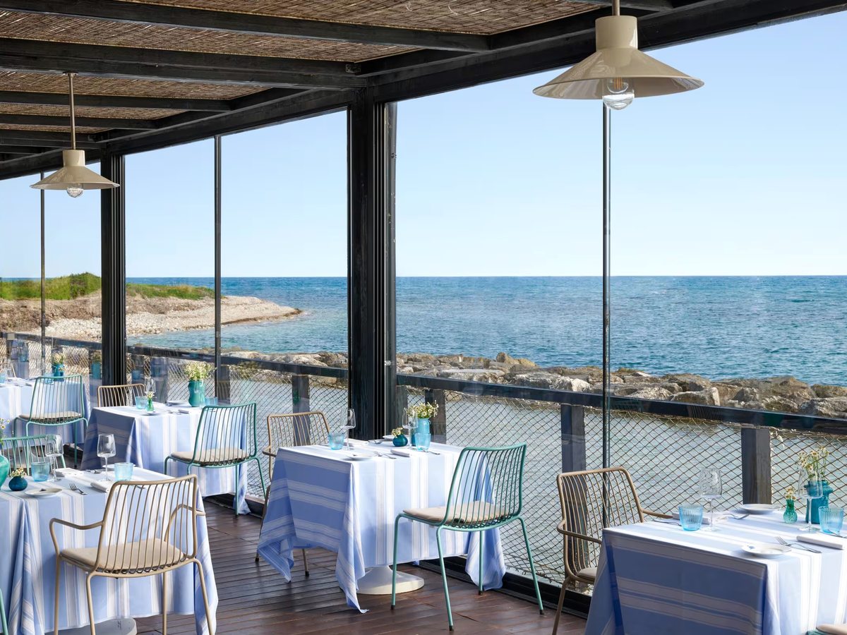 Covered seafront dining terrace with blue-and-white striped tablecloths, pale metal chairs and hanging lamps, open to a rocky shore and blue sea.
