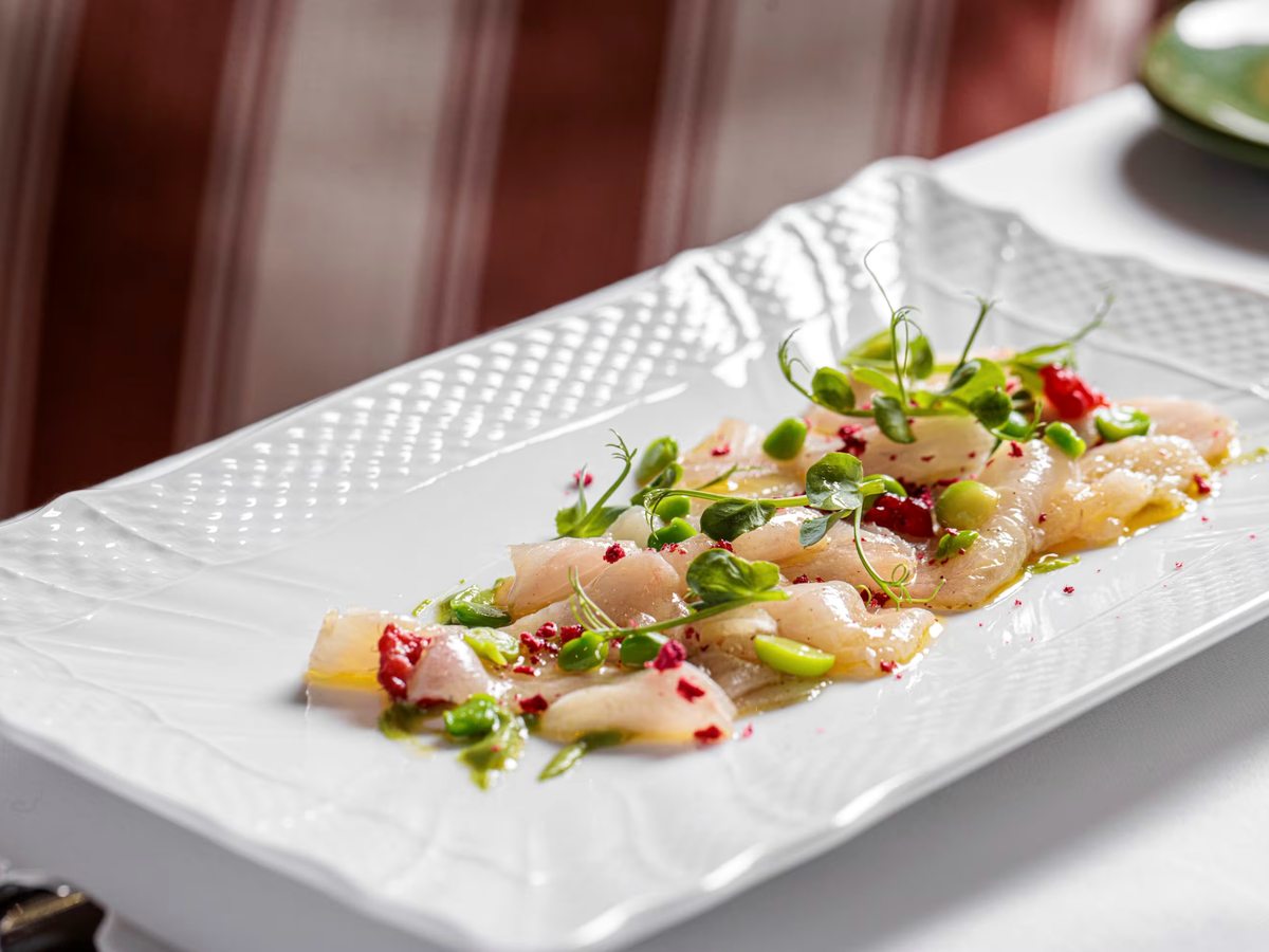 Thin slices of raw white fish on a long white platter, dressed with olive oil, broad beans, micro leaves and flecks of red.