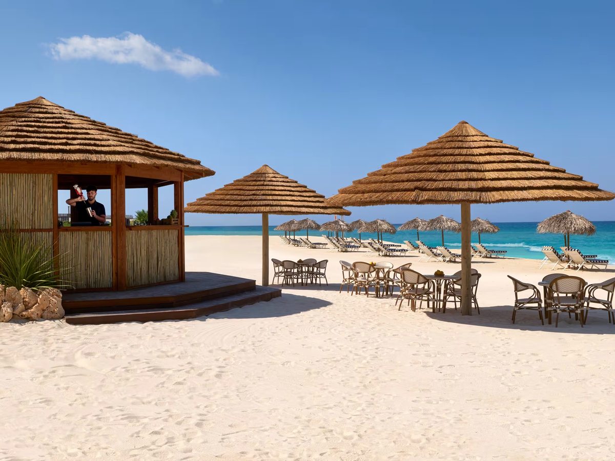 Thatched timber beach bar with a barman at the counter, and cafe tables and thatched parasols on white sand beside a turquoise sea.
