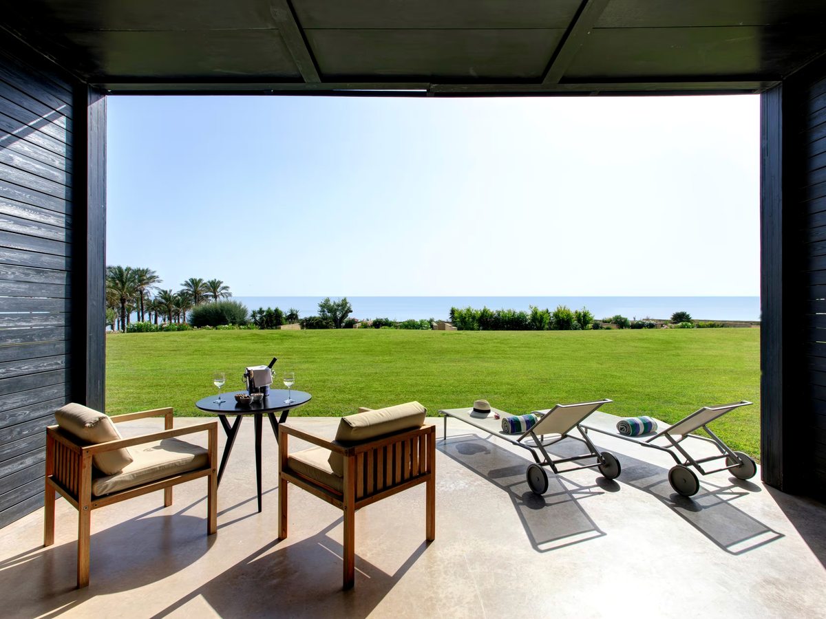 Shaded room terrace with wooden armchairs, a round table and two sun loungers, opening level onto a wide green lawn with palms and the sea beyond.