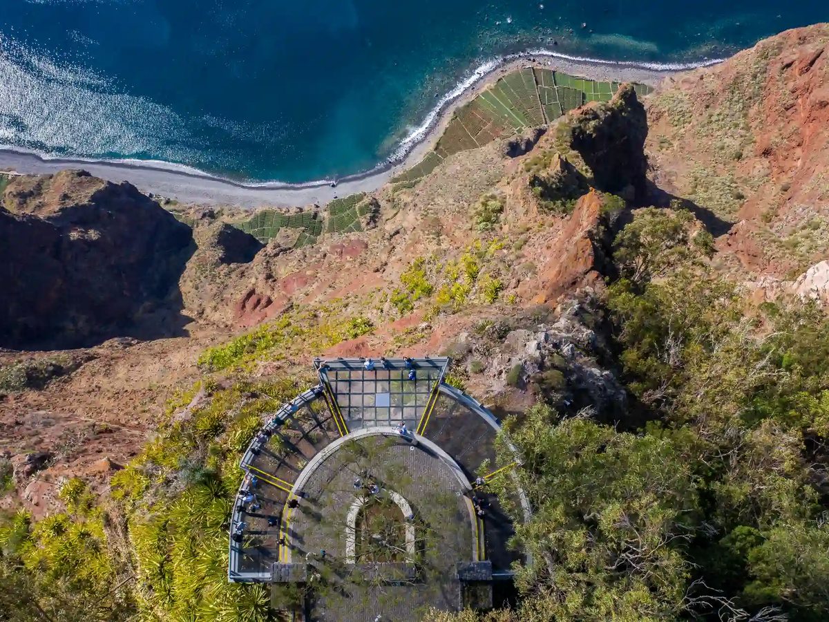 A semicircular glass viewing platform on a cliff edge, with terraced fields, a pebble shore and blue sea far below.