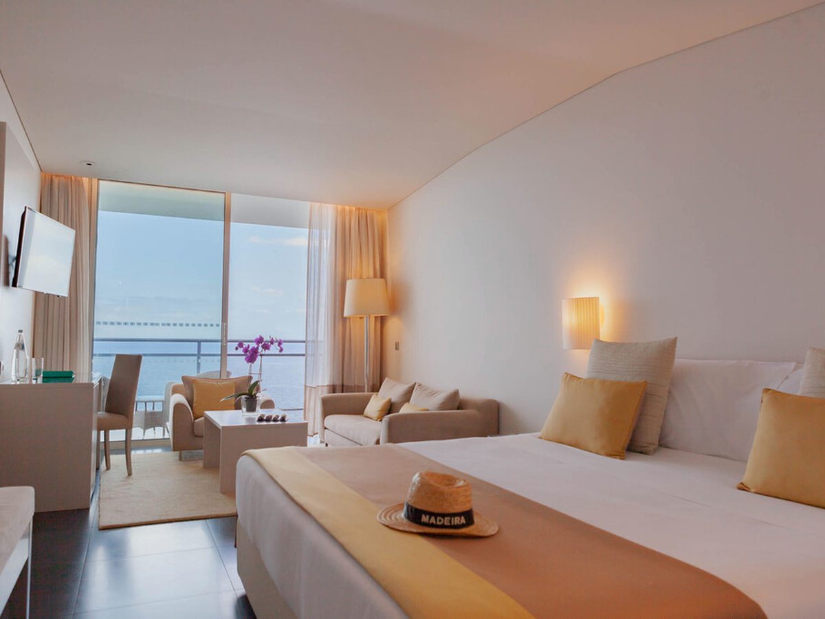 A double room in cream and gold with a wide bed, a sofa and armchair by the window and a balcony looking straight out to the sea.
