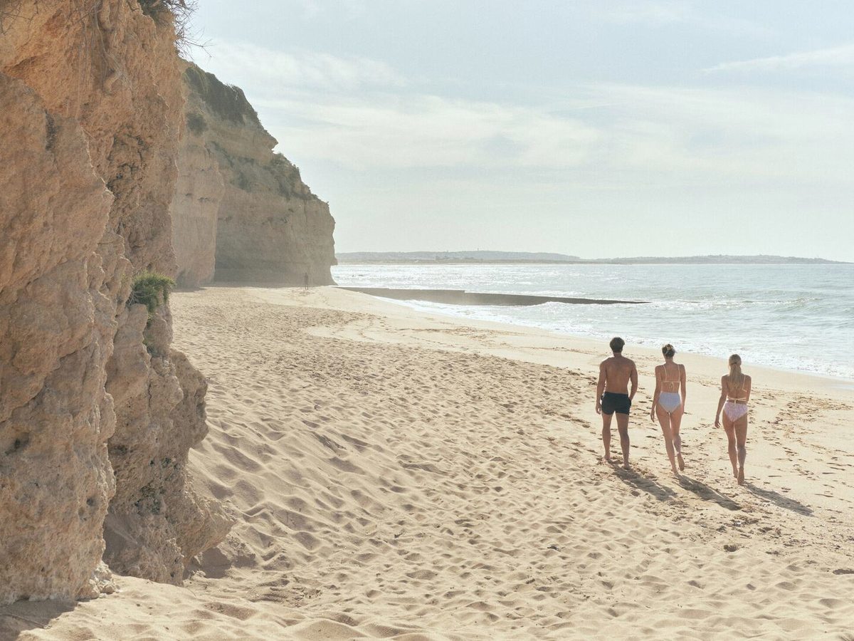A wide stretch of soft sand at the foot of a tall ochre cliff, with three people walking towards the sea in hazy sunlight.