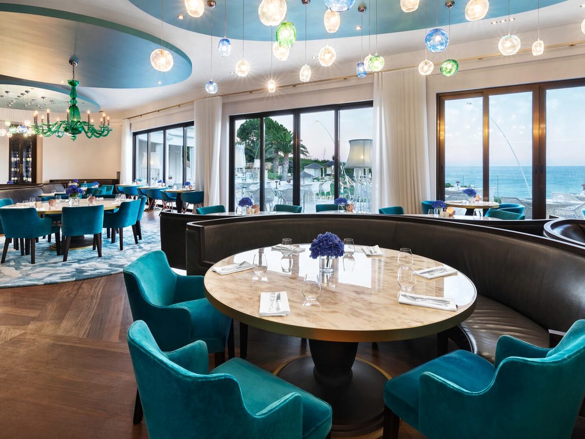 A restaurant interior with teal velvet armchairs around marble tables, a curved leather banquette, coloured glass pendant lights and full-height windows to the sea.