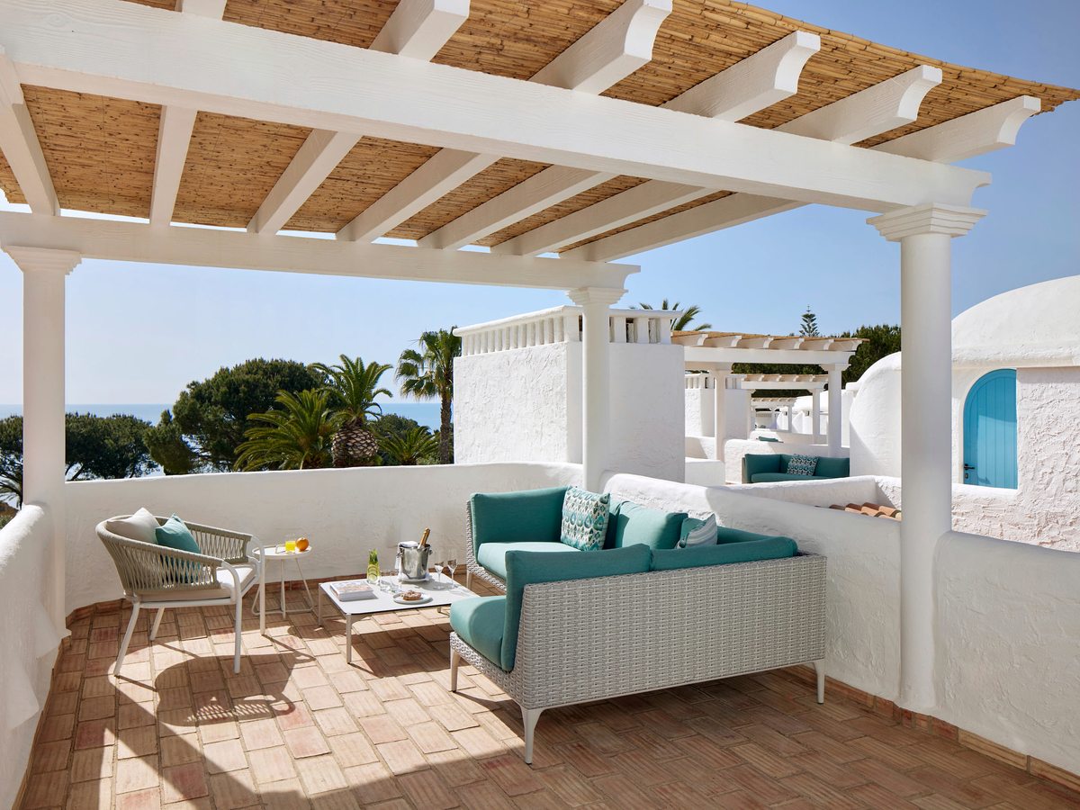 A whitewashed rooftop terrace under a timber pergola, with a teal wicker sofa and armchair on terracotta tiles, and domed white rooftops and palms beyond.