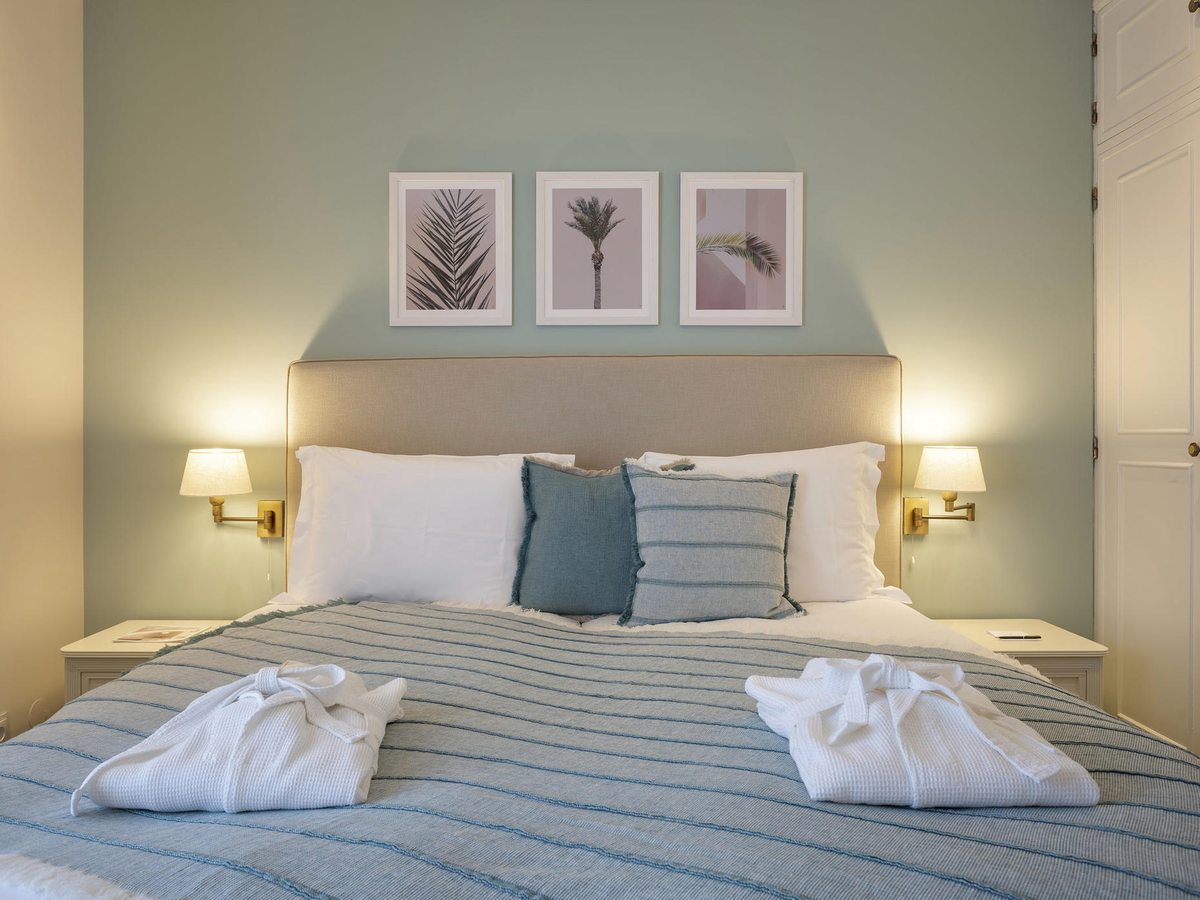 A bedroom with a pale green wall, an upholstered headboard, three framed palm prints, brass wall lamps and a striped blue throw across the bed.
