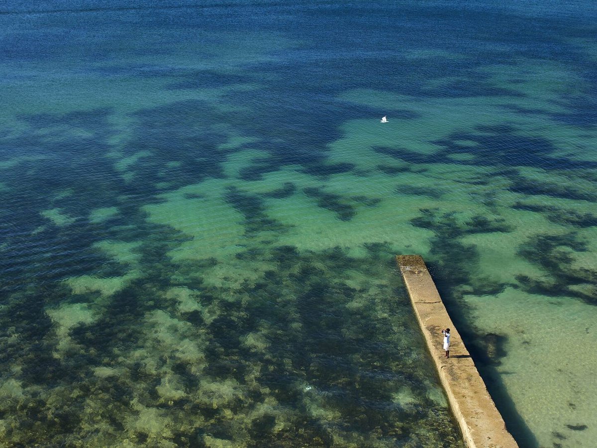 Clear green and blue water over patches of rock, with a narrow concrete jetty running out from the shore and a person standing at its end.