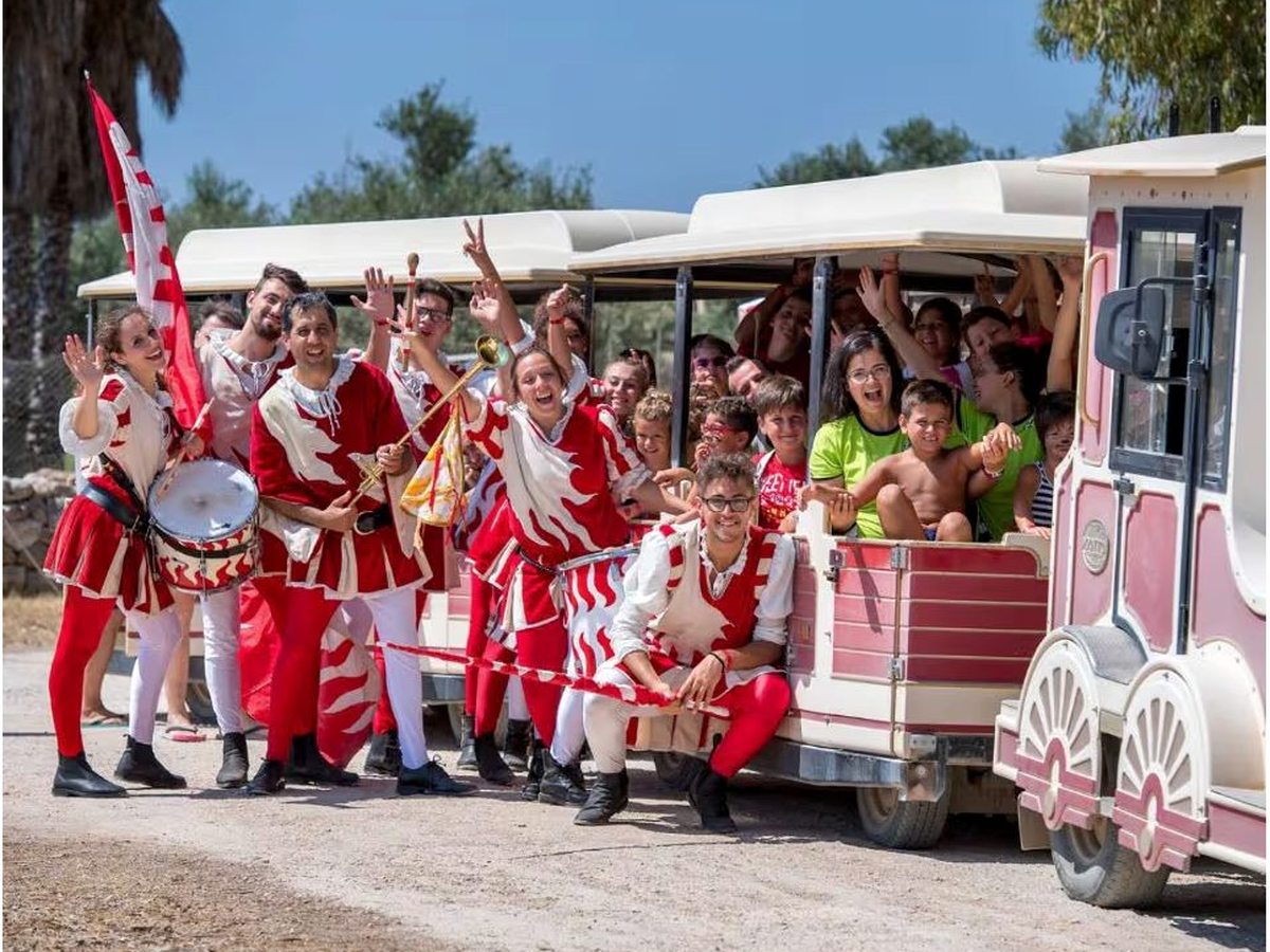 A pink and cream road train packed with waving children and parents, with the resort's entertainment team in red and white costumes standing alongside it, one of them carrying a drum.