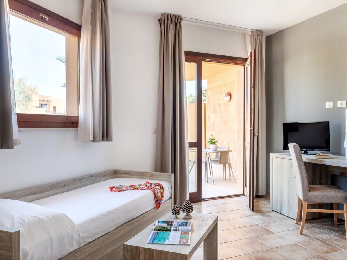 A living room with two single beds along the wall, a low wooden coffee table and a desk with a television, beside open doors onto a furnished terrace with a table and chairs.