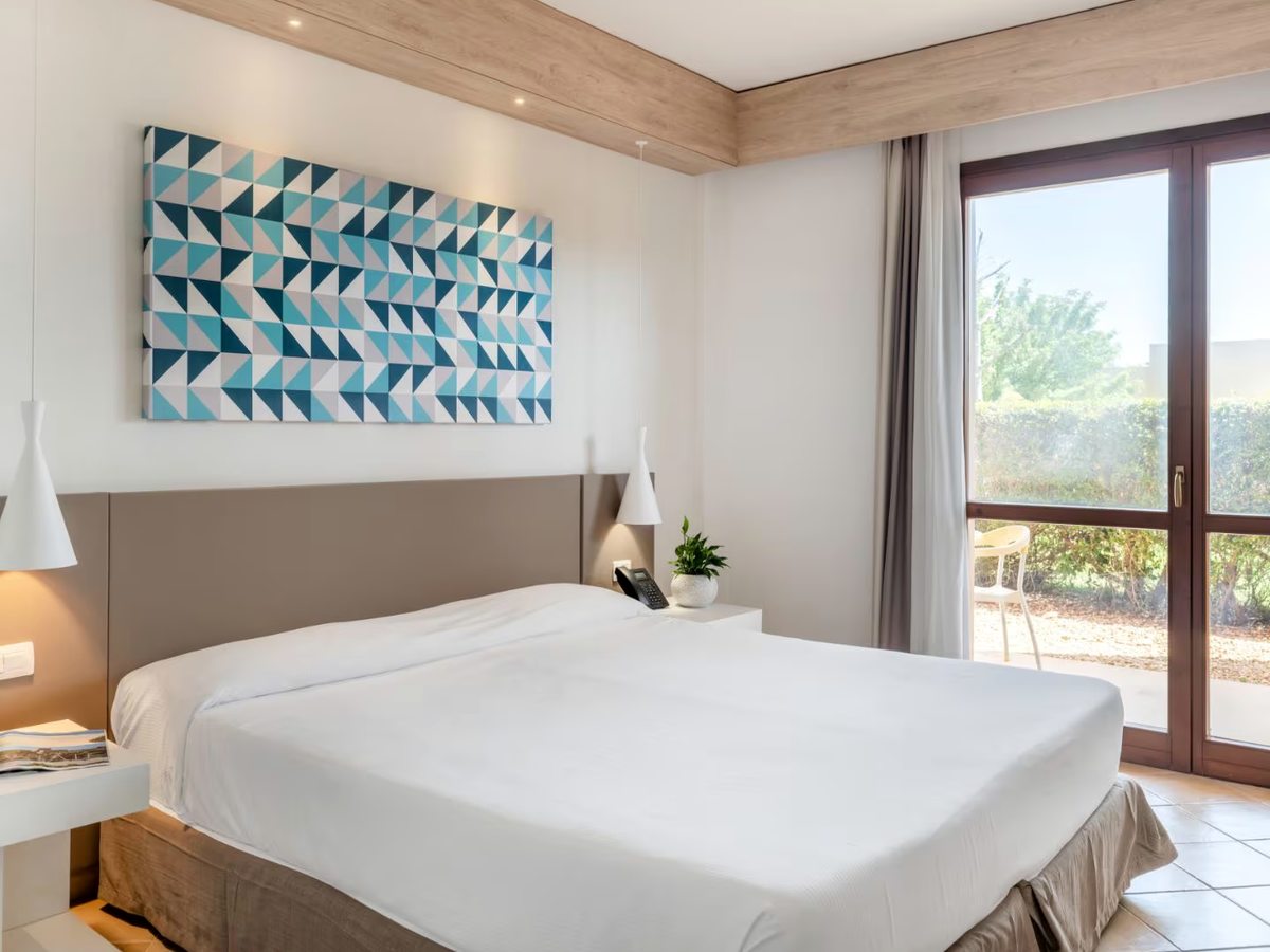 A double bedroom with a taupe upholstered headboard and a blue and white geometric artwork above it, next to full-height glazed doors opening onto a garden terrace with a chair and a clipped hedge.