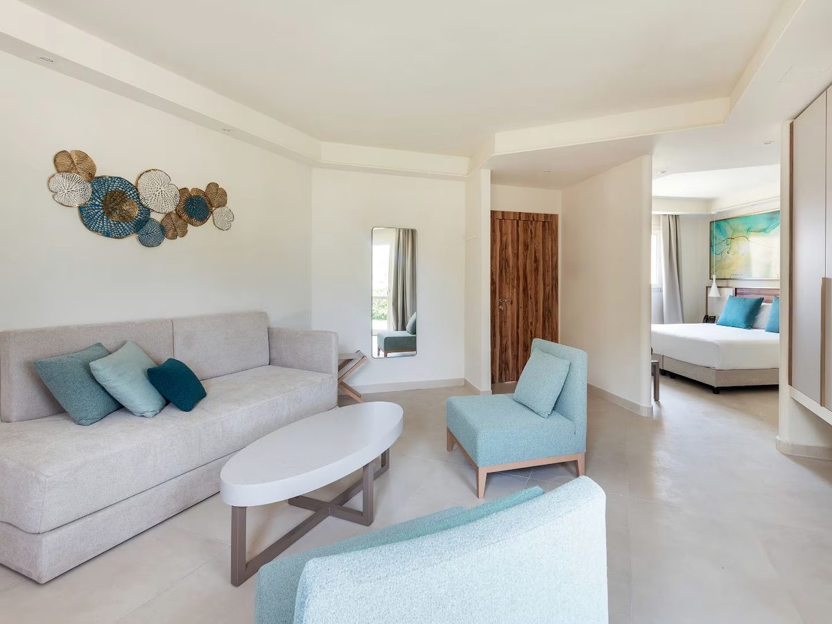 A large suite living area with a long pale sofa, two light blue armchairs and an oval coffee table, with a wide opening through to a double bedroom beyond.