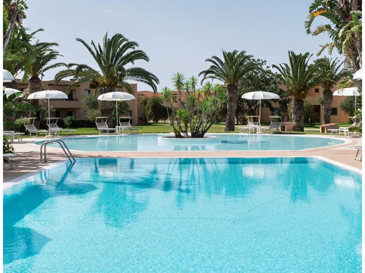A large outdoor pool with a shallower upper basin beside it, set around a planted island of yuccas and edged with white parasols, sun loungers, palms and low ochre buildings.