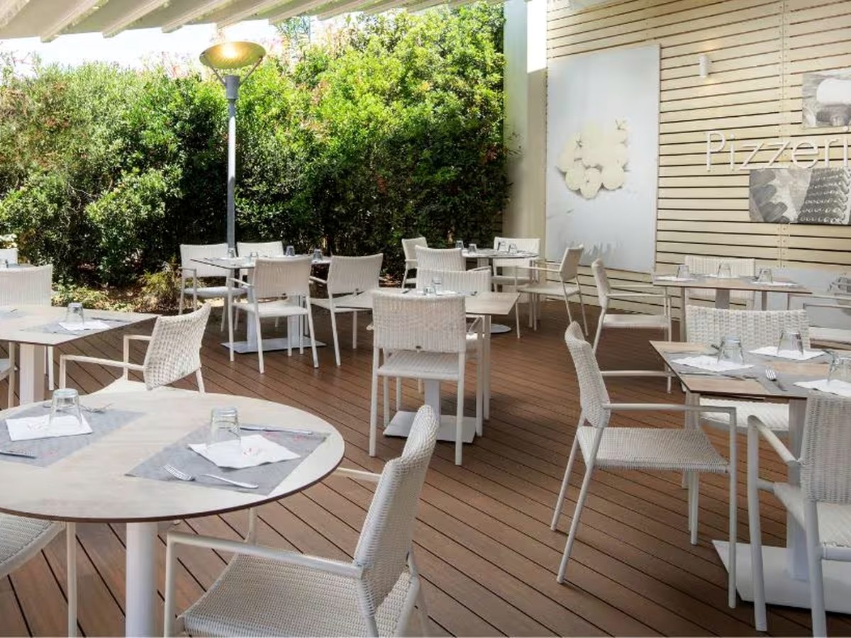 A decked outdoor restaurant terrace with white tables and woven chairs against a tall hedge, with a Pizzeria sign on the timber wall.