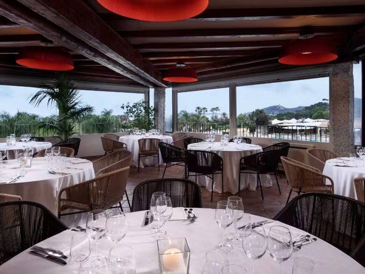 A covered upper terrace with round tables in white linen, red pendant lampshades and open sides looking over the pool and the hills beyond.