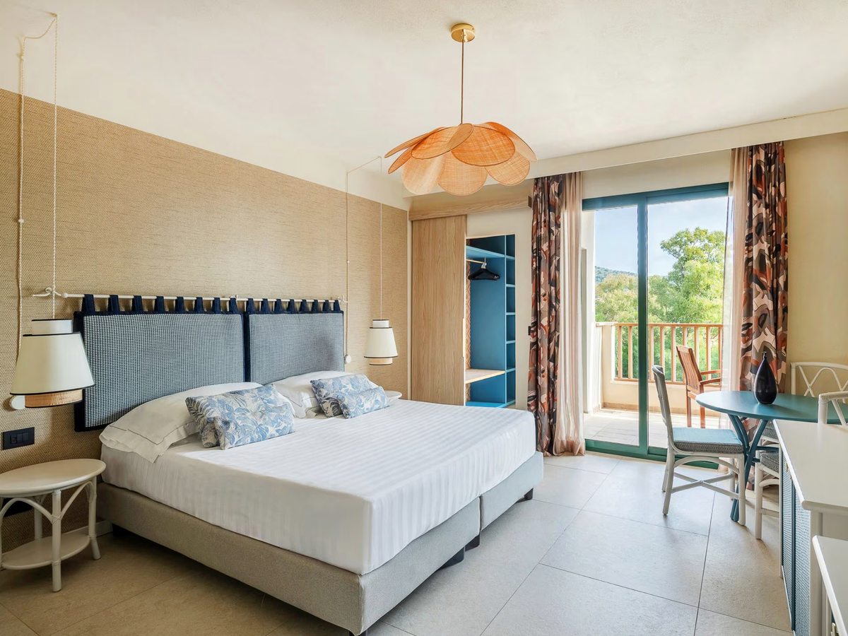 A double bed with a blue quilted headboard, a rattan pendant light, a walk-in wardrobe and doors onto a balcony with a table and chairs.
