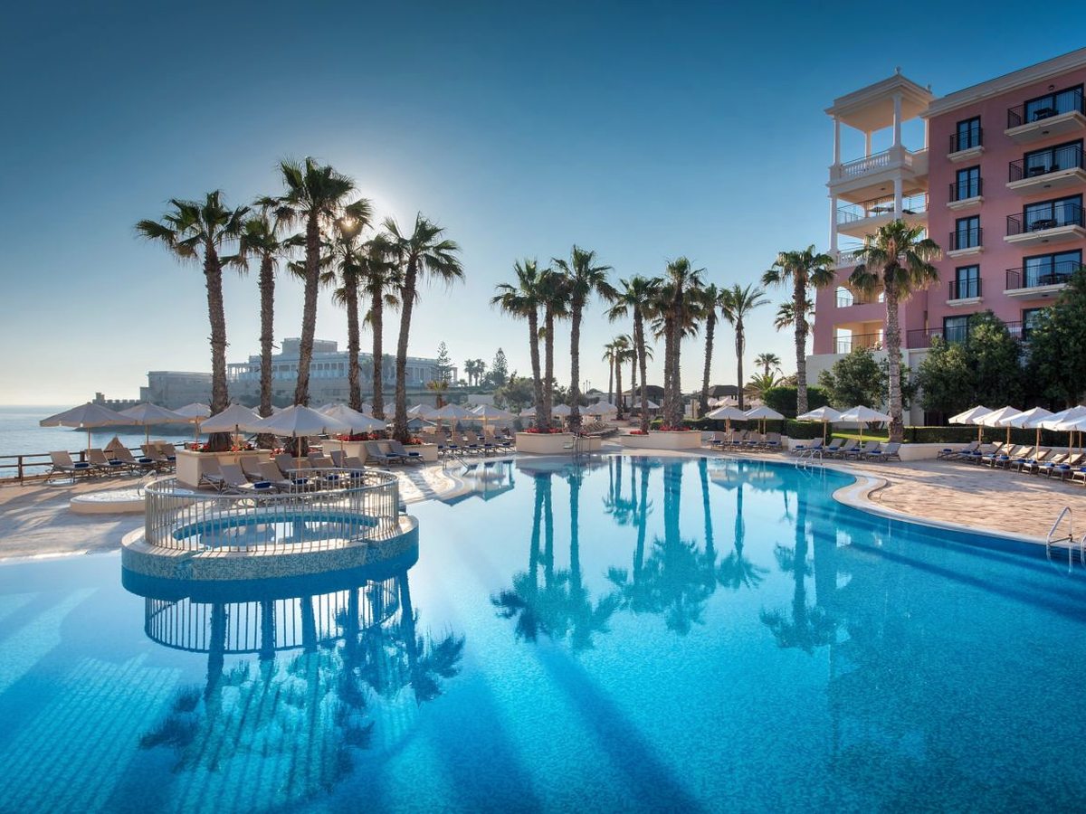 A large curved outdoor pool edged with palms and parasols beside the pink hotel building, with a round whirlpool at one end and the sea beyond.