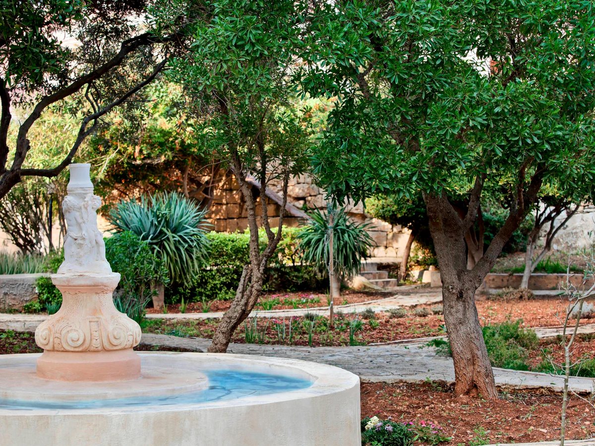 A carved stone fountain with a small statue in a walled garden of mature olive trees, agaves and planted beds crossed by stone paths.
