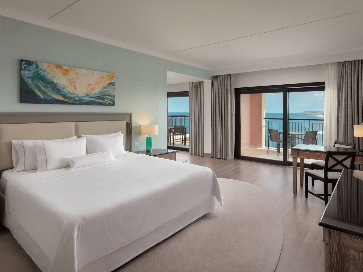 A hotel bedroom with a king bed, a seascape painting above the headboard, a desk and chair, and sliding doors onto a balcony with the sea beyond.