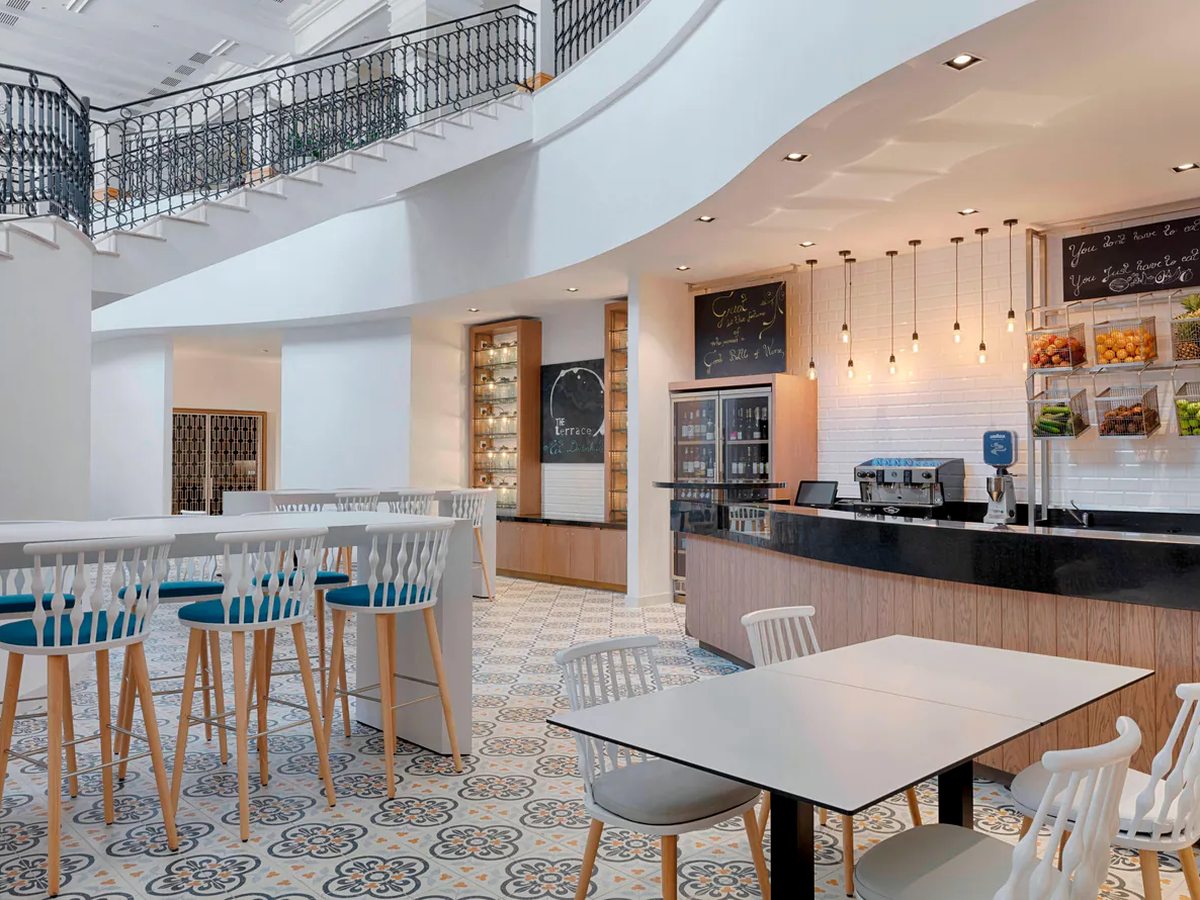 A cafe counter with an espresso machine, wire baskets of fruit and chalkboard menus, white bar stools at a high table and patterned tiled flooring, below a curved balcony with wrought-iron railings.