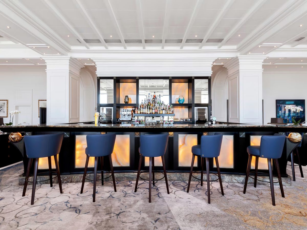 A long marble-topped bar with backlit onyx panels and blue leather stools, spirit bottles on shelves behind and a white coffered ceiling above a patterned carpet.