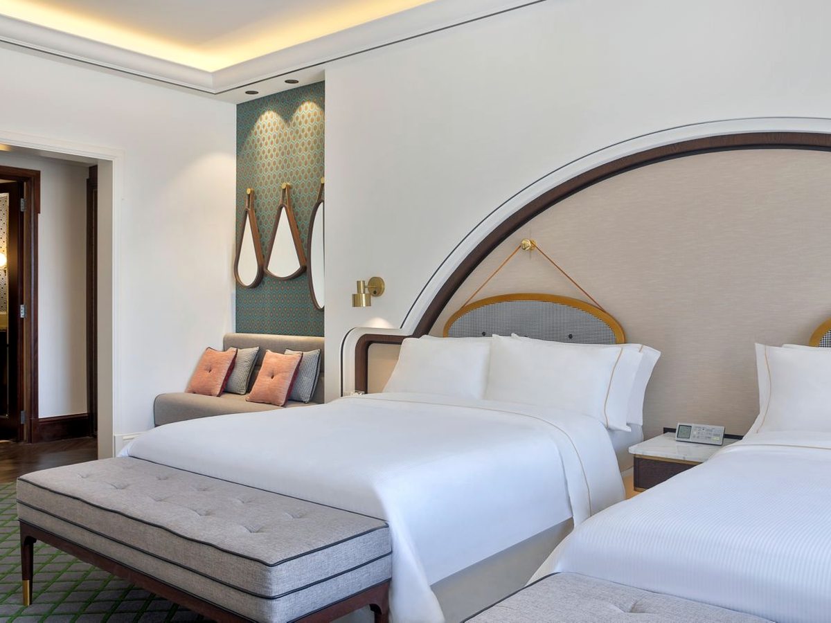 A guest room with two white-linen beds side by side under a wide arched padded headboard, a grey upholstered bench at the foot, a small sofa against a patterned teal panel and a doorway through to the bathroom.