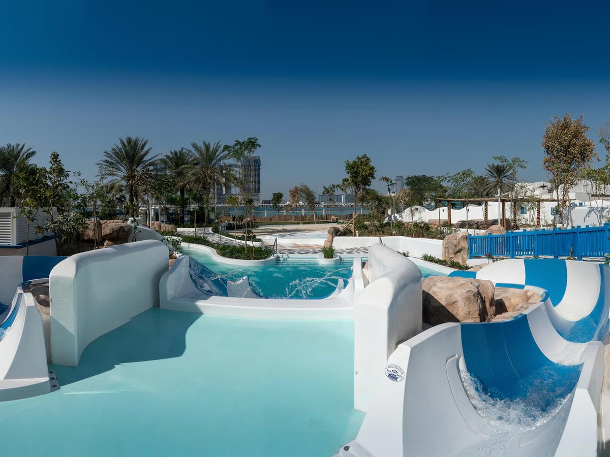 White and blue waterpark chutes curving down into a shallow turquoise pool with rock edging, with palms, planting and the sea and city skyline behind under a deep blue sky.