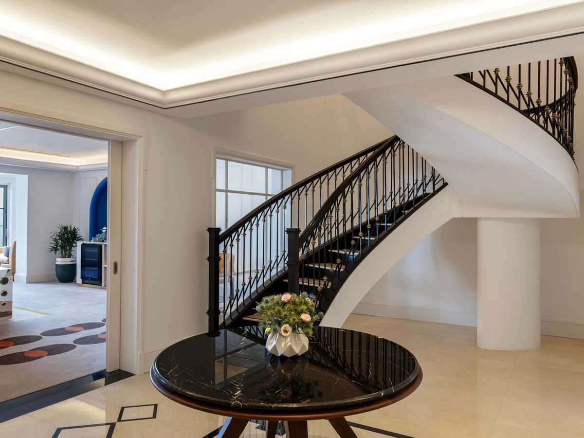 A cream marble entrance hall with an inlaid border, a sweeping staircase with a black ironwork balustrade curving up to the floor above, and a round black marble table with a flower arrangement in the foreground.