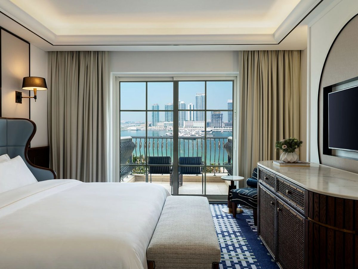 A guest room with a king bed and a long bench, a dark rattan-fronted dresser and television, and sliding glass doors onto a balcony with two chairs facing water and a line of high-rise towers.