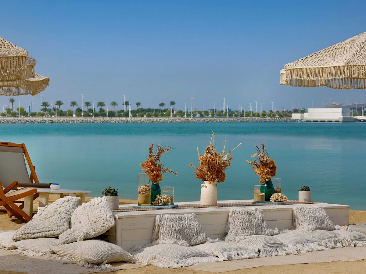 Low white cushioned bench seating with bolsters and floor cushions on the sand, with dried floral arrangements in ceramic vases along the bench top and calm turquoise water directly behind.