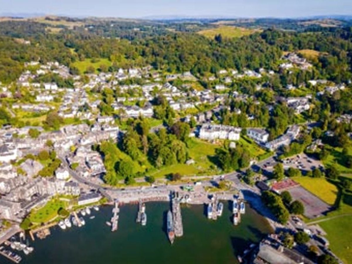 An aerial view of Bowness-on-Windermere, with its piers and cruise boats on the lake, the grey stone town behind and wooded hills rising beyond.