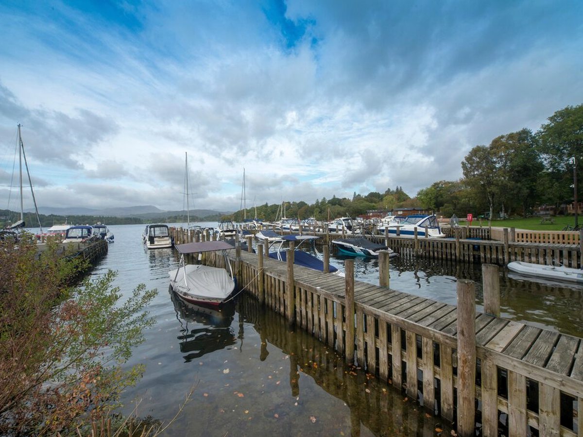 Timber pontoons reaching out into Lake Windermere with motorboats and yachts moored along them, wooded shoreline to the right and low fells across the water.