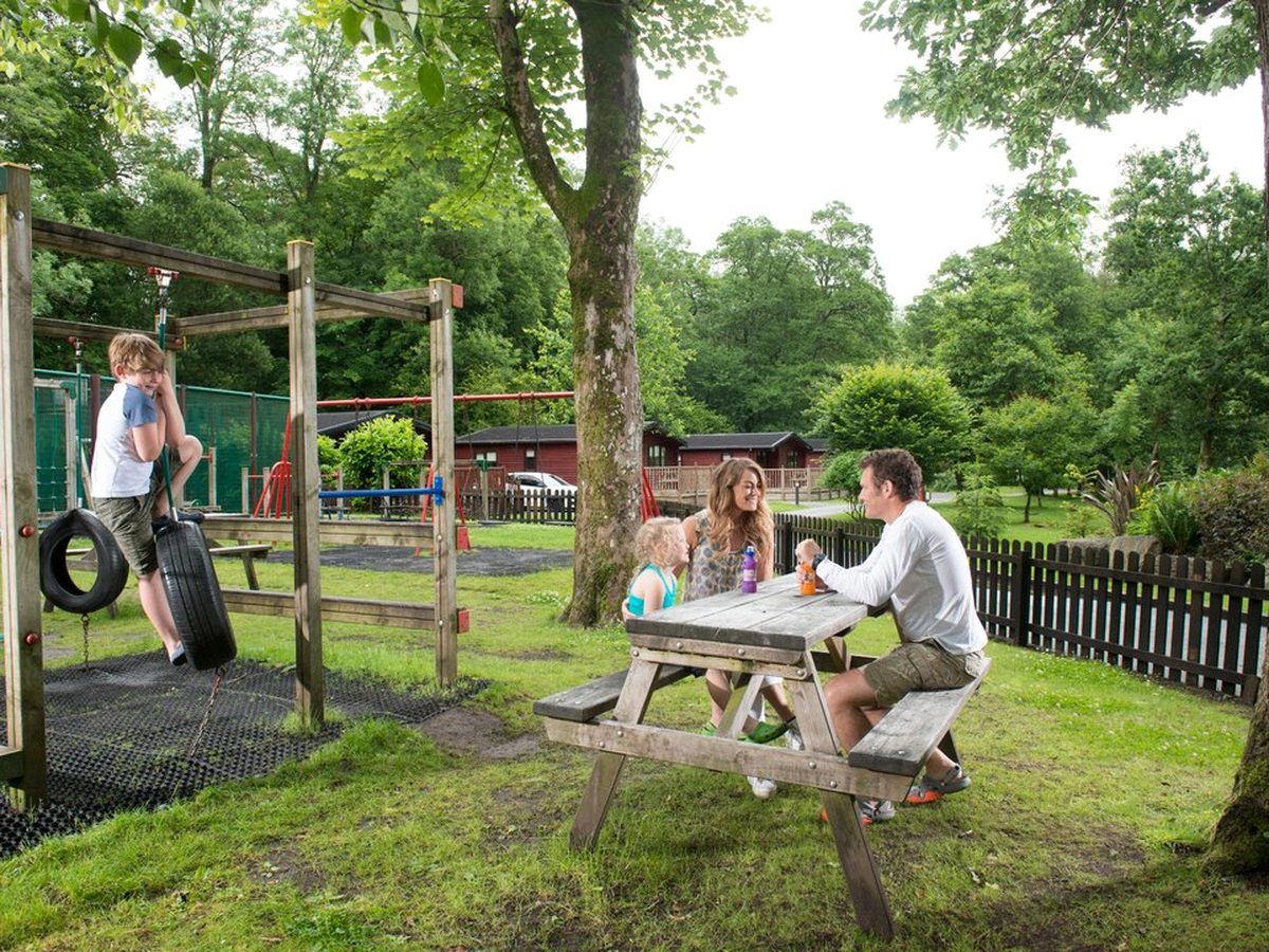 A timber adventure playground under trees, with tyre swings, climbing frames and a slide over green safety surfacing and picnic tables on the grass alongside.