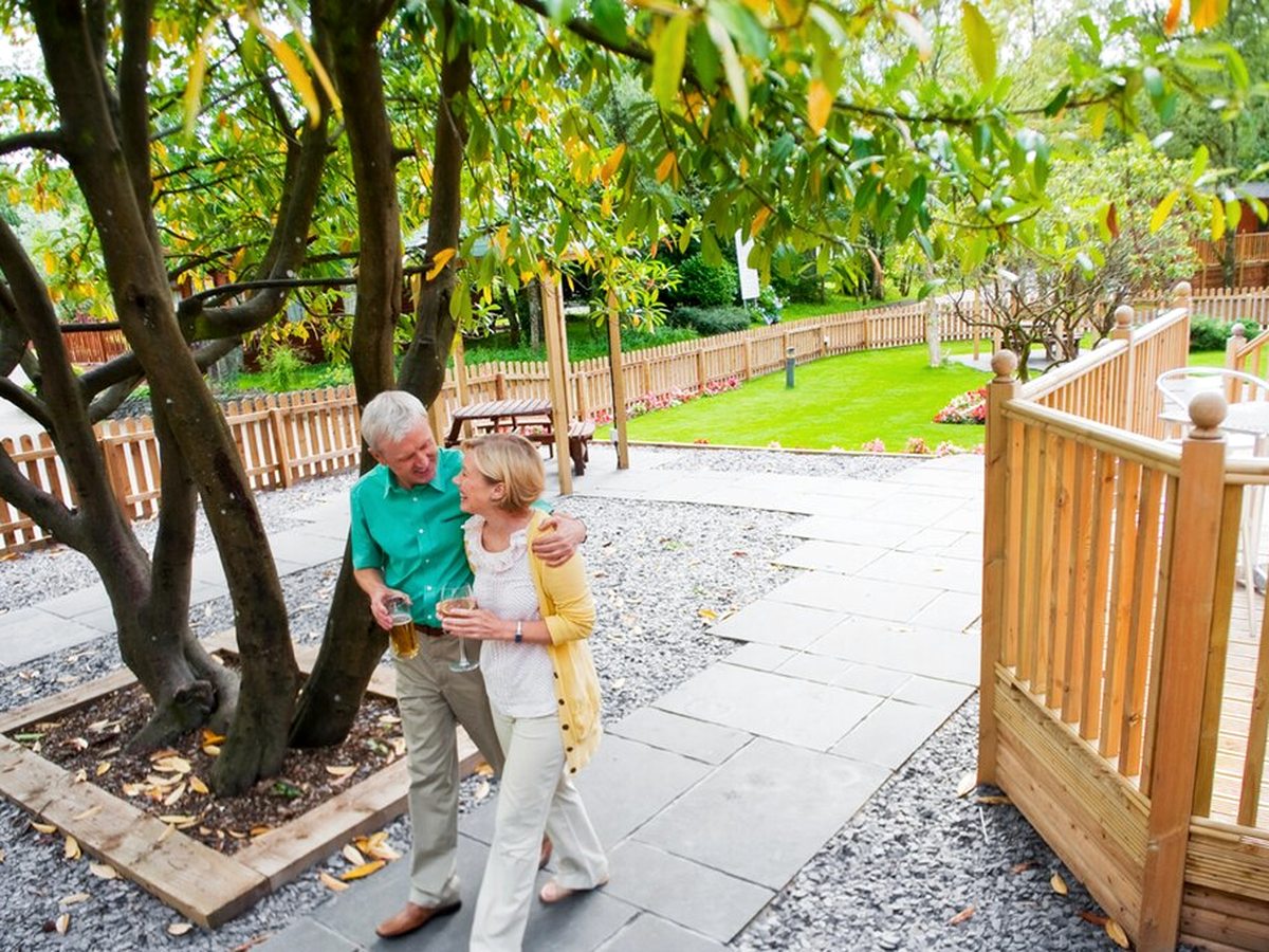 A paved footpath curving between gravel beds and low shrubs beneath a mature tree, with a timber handrail alongside and an older couple walking down it.