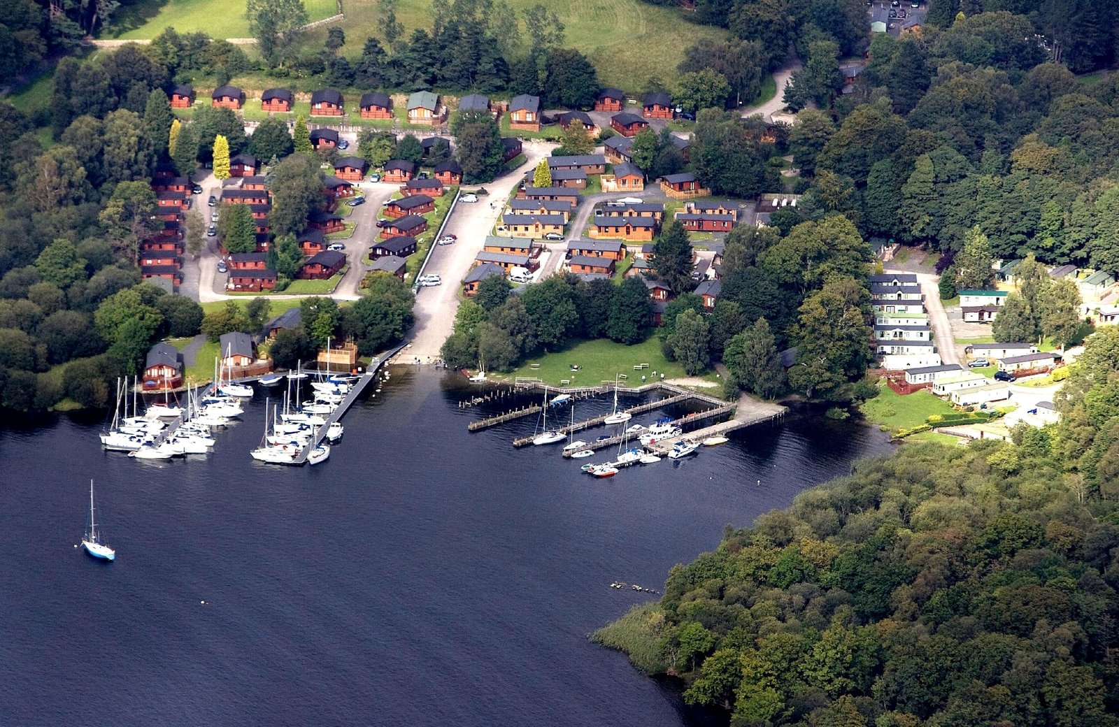 An aerial view of White Cross Bay Holiday Park on the wooded eastern shore of Lake Windermere, with timber lodges and caravans among the trees and yachts moored along the marina pontoons below.