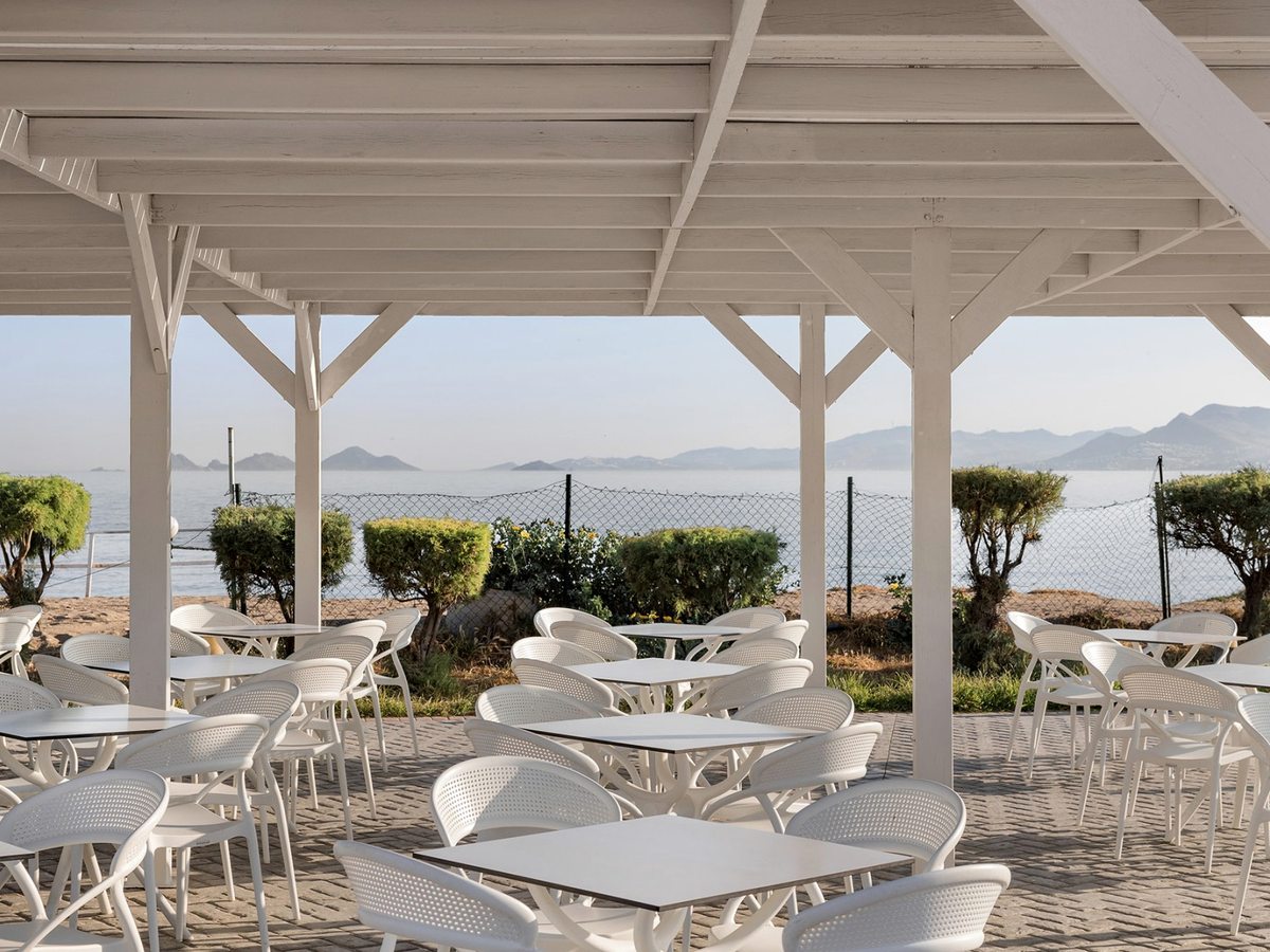 White timber pergola over white tables and chairs on a paved terrace, with clipped shrubs, the shoreline and distant hills beyond.
