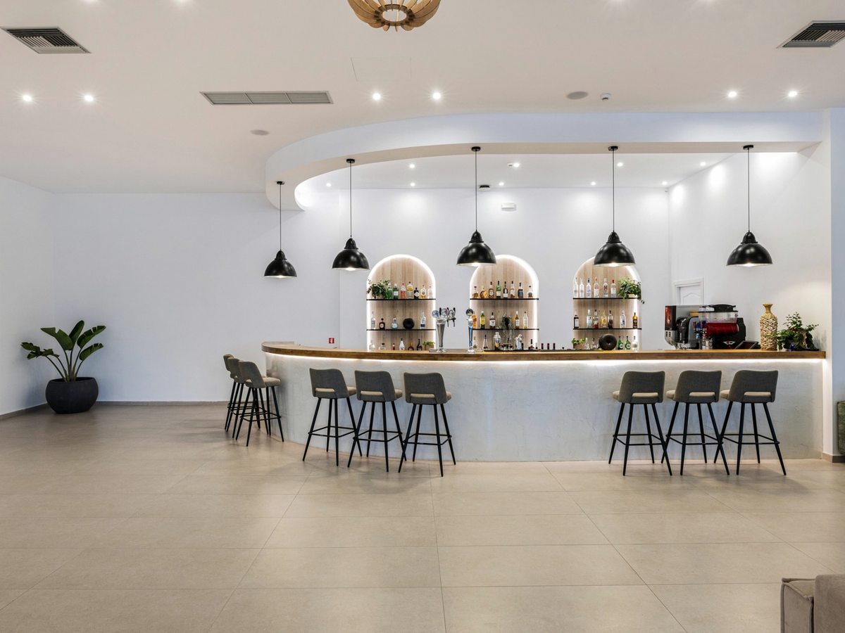 Curved bar with a timber counter, grey bar stools, arched shelving of spirits, a coffee machine and black pendant lights in a pale-tiled room.