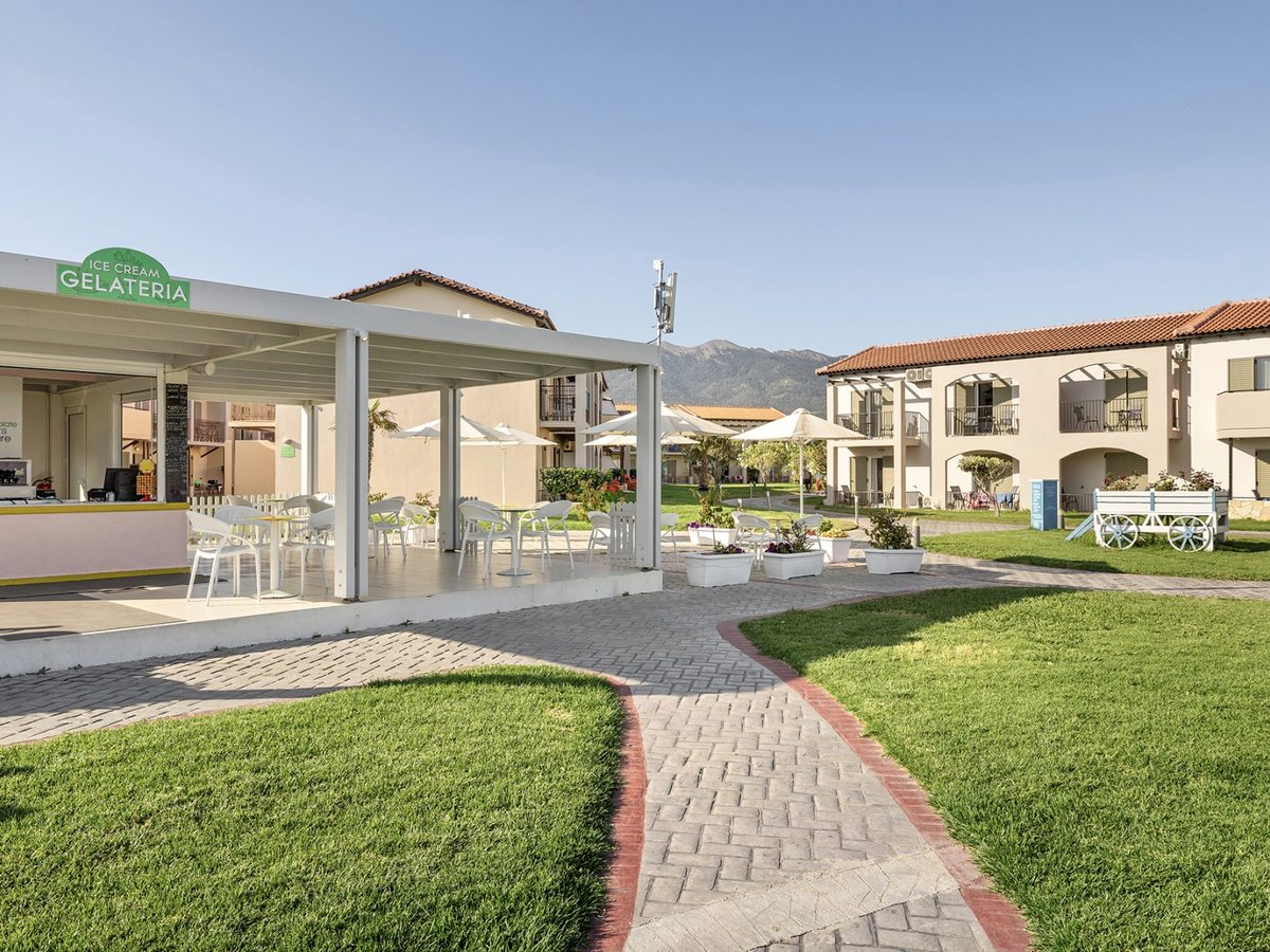 Small open-fronted ice cream kiosk signed Gelateria, with a white pergola over tables and chairs, lawns and brick paths around it and room blocks and mountains behind.