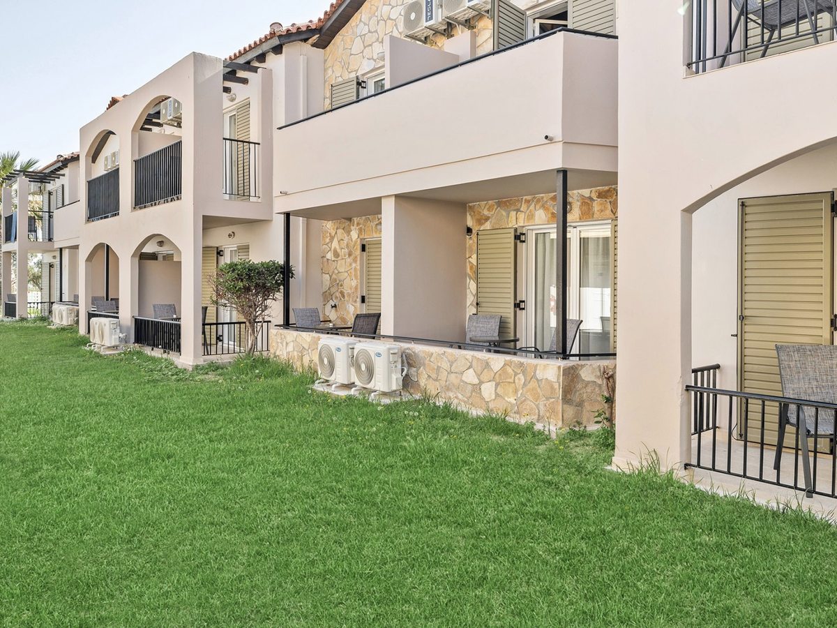 Two-storey stone-clad accommodation block with covered ground-floor patios and railed first-floor balconies, set behind a mown lawn.