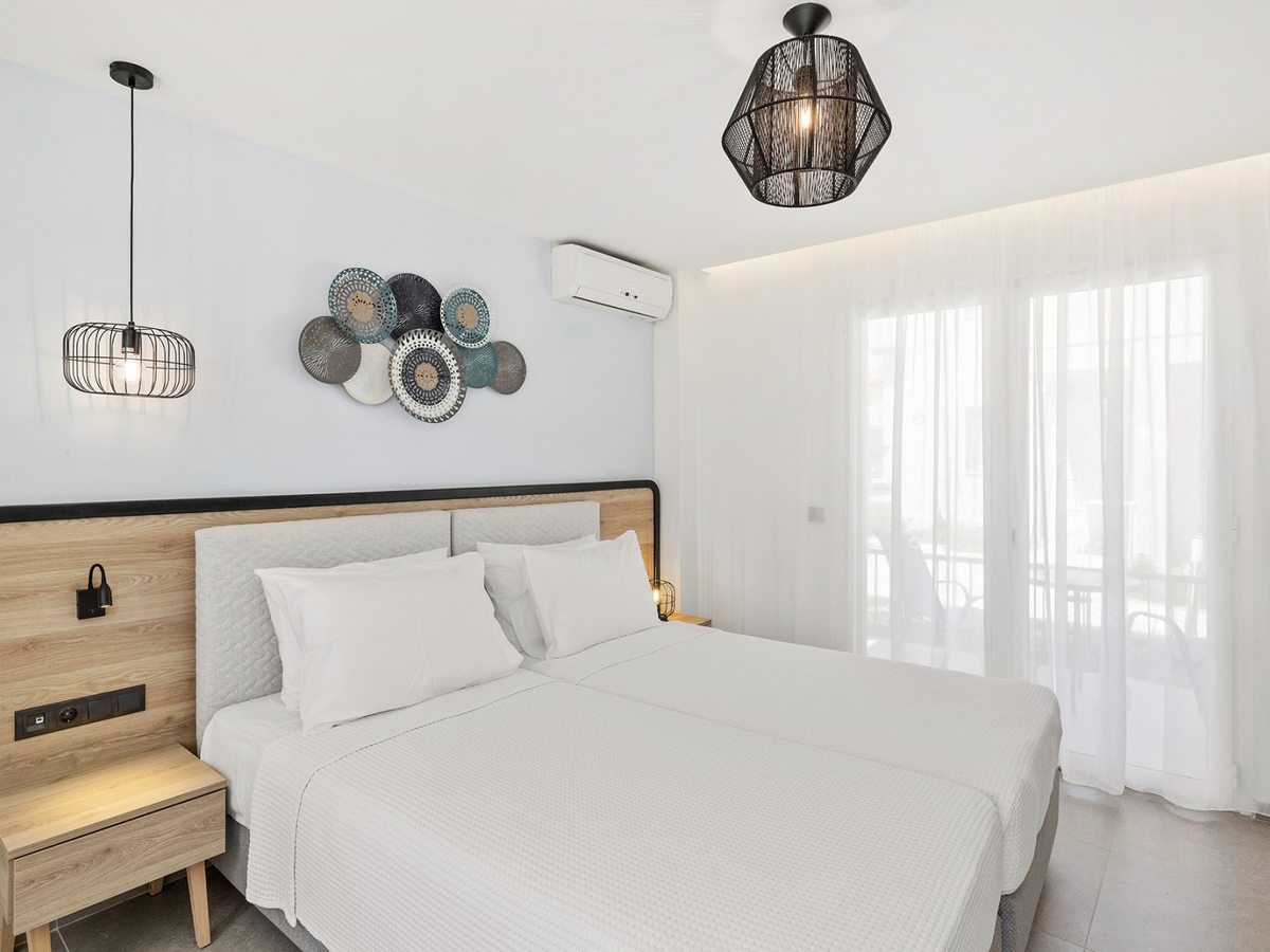 Double bed with a wood and upholstered headboard, plate-style wall art, an air conditioning unit and pendant lights, with curtained balcony doors alongside.