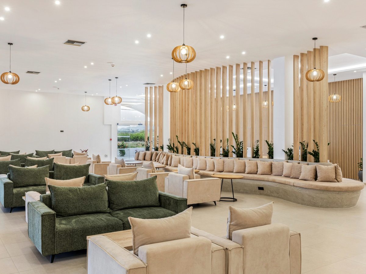 Bright open-plan lobby lounge with green and cream sofas and armchairs, a long curved cushioned banquette, timber-slat screens planted with greenery and woven globe pendant lights.