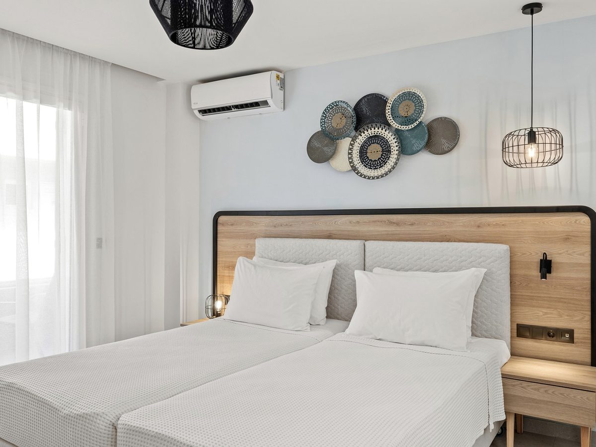 Two single beds pushed together with white bedding, under a wood and upholstered headboard with plate-style wall art and a pendant light, beside a curtained window.