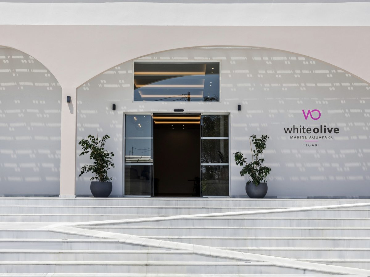 White stucco entrance under an arch, with sliding glass doors, the White Olive Marine Aquapark Tigaki sign, two potted plants and a wide run of marble steps.